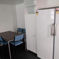 A small dining table surrounded by blue chairs, next to a large fridge and freezer and some cupboards. 