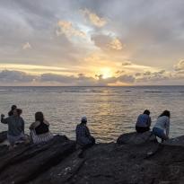A group of people sit on the coast admiring the sunset over the ocean