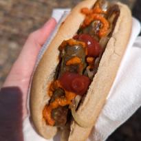 Hand with napkin holding a bun with mini brown sausages (Bratwurst) inside, with an orange chili sauce and ketchup squeezed on top