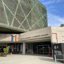 A glass and stone building set against a blue sky, there is a pride flag and anti-torture display in front.