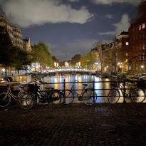 Bikes sitting by the rail of a canal in Amsterdam at night. The edges of a bridge in the distance are lit up, lighting the water and the scenery around.