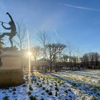 A statue in the Luxembourg Gardens with the sun behind it and green grass peeking out from under the snow.