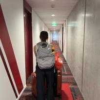 This photo features a girl with a purple backpack and two orange suitcases. She's rolling the suitcases down a hallway that has red carpet and red wall features.