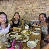 In this photo, there are four people sitting at a table with four plates of Thai food. They're all smiling brightly as the pose for the selfie. There is brick wall behind them.