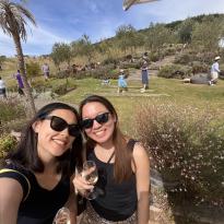 In this photo, two people pose for a selfie in an outdoor space at a vineyard. They both wear black sunglasses, black shirts, patterned skirts, and hold wine glasses in their hands. In the background, there are wooden steps, trees, shrubs, benches, bean bags, and people. The sky has a few clouds.