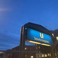A blue U-Bahn station sign reading "Senefelderplatz" with a sunset in the background