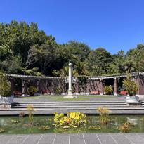 In this photo, there is a sculpture with a fountain in front. In the fountain, there are several lily pads. Between the sculpture and the fountain there are stairs. There are bricks behind the sculpture and fountain to enclose the area from the tall trees that are in the background. 