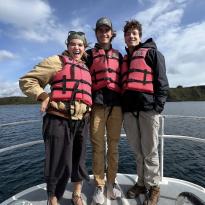 Three friends posed at the helm of a boat, standing. Lifejackets on and smiling.