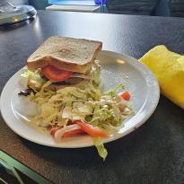 A sizeable beef burger with melted cheddar and young + mild cheese topped with sliced tomato, iceberg lettuce, sauced with barbeque. Accompanied with leftover lettuce and tomato for a side salad.