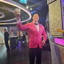 wax statue of the rock wearing a pink suit and black bowtie. he is also bald.