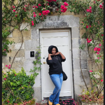 The author of this blog is posing in front of a white door that is the backdrop of a pink floral arch in Maastricht 