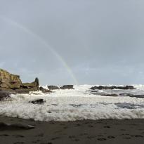 A rainbow over a seafoam covered black sand coast.