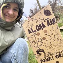A selfie of Pono with a cardboard plant protector that has a drawing and the words "Aloha ʻĀina" and "Mauri Ora".