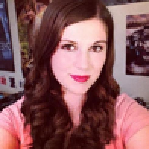 Smiling woman with long brown hair and pink top, colorful posters in background. This alt text was added with Al; accuracy may vary.