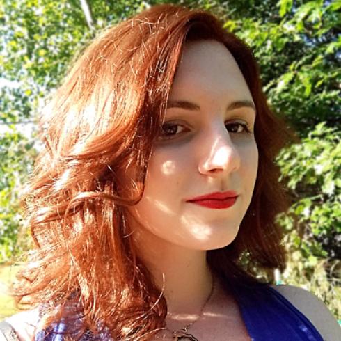 Red-haired person smiling outdoors, green foliage backdrop. This alt text was added with Al; accuracy may vary.