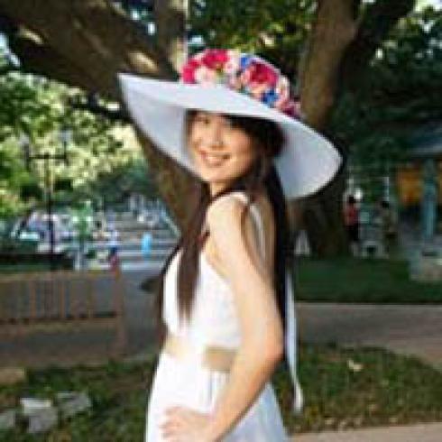 Smiling woman outdoors wearing a white hat with flowers under a large tree. This alt text was added with Al; accuracy may vary.