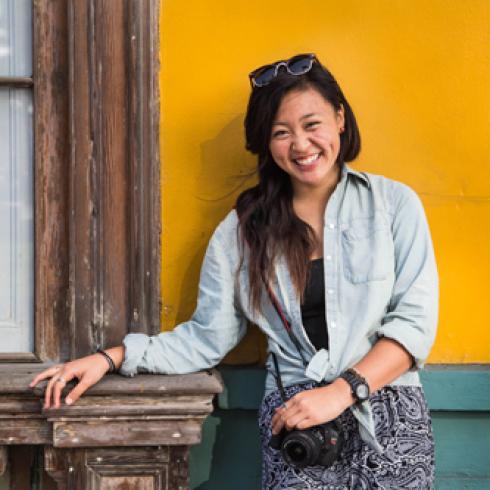 Smiling woman in denim by a yellow wall, holding a camera. This alt text was added with Al; accuracy may vary.