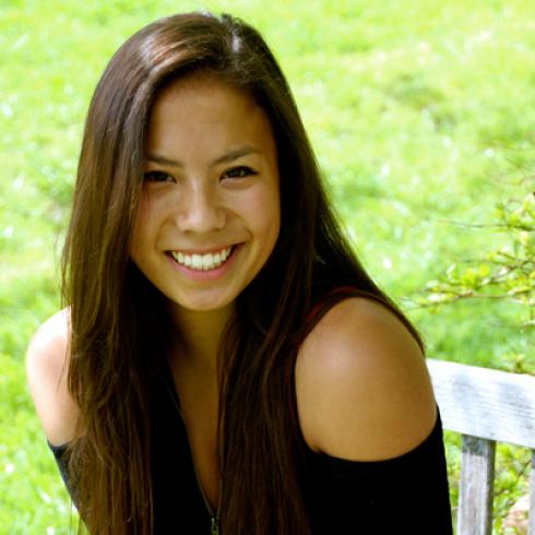 Smiling woman with long hair sitting on a bench in a grassy area. This alt text was added with Al; accuracy may vary.