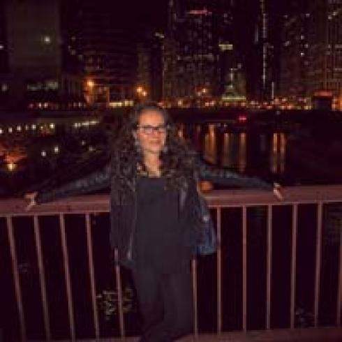 Smiling person in a black outfit on a bridge at night, city lights in the background. This alt text was added with Al; accuracy may vary.