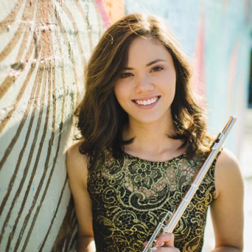 Young woman smiling, holding a flute, standing against a patterned wall. This alt text was added with Al; accuracy may vary.