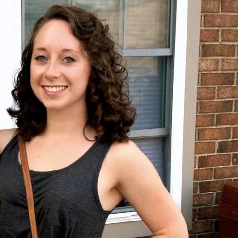 Smiling person with curly hair stands outdoors near a brick wall and window. This alt text was added with Al; accuracy may vary.