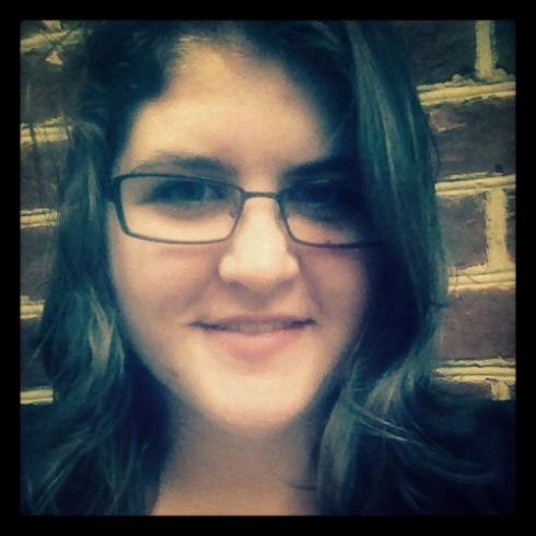 Smiling person with dark hair and glasses, brick wall background. This alt text was added with Al; accuracy may vary.