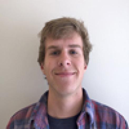 Young man smiling against a plain background, wearing a plaid shirt. This alt text was added with Al; accuracy may vary.