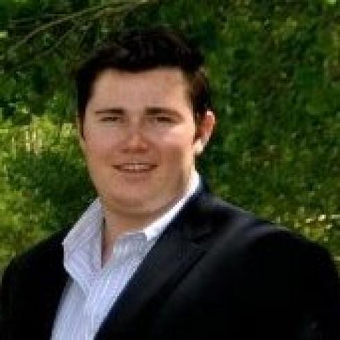 Smiling person outdoors, wearing a dark blazer with greenery in the background. This alt text was added with Al; accuracy may vary.