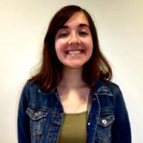 Circular window view of a statue-topped building, overcast sky.

Ruins and historic buildings in a sunlit courtyard.

Looking down a spiral stone staircase, curved steps.

Person smiling, wearing a denim jacket against a plain background. This alt text was added with Al; accuracy may vary.