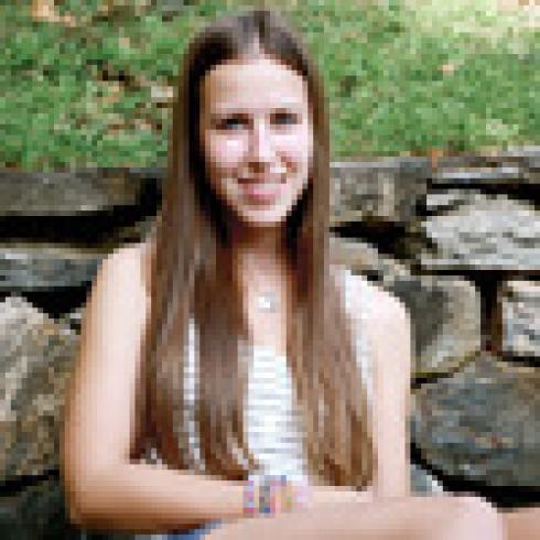 Young woman with long hair smiling, sitting on stone steps outdoors. This alt text was added with Al; accuracy may vary.