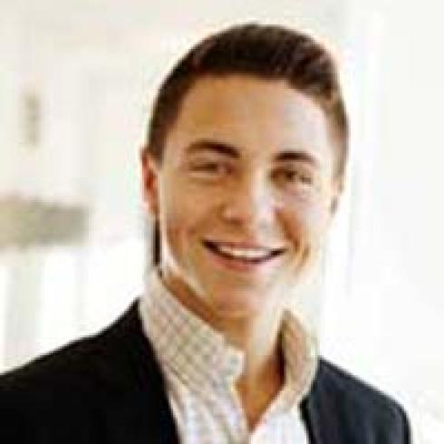 Smiling person with short hair in a well-lit room, wearing a suit and checkered shirt. This alt text was added with Al; accuracy may vary.