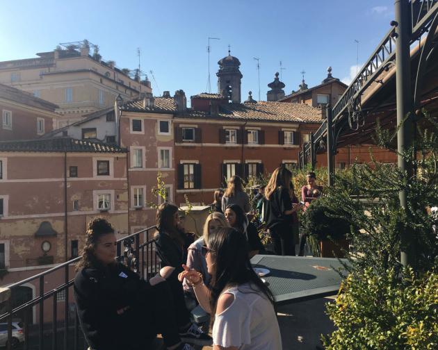 Students sit at cafe tables on a rooftop terrace. In the background are several building rooftops and a blue sky.
