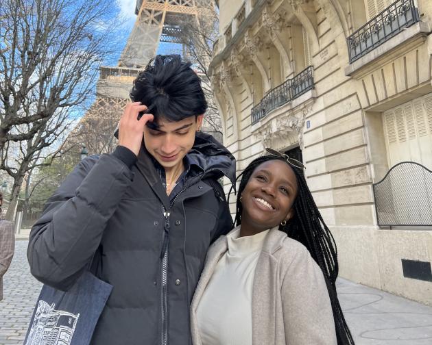 Two students, one taller than the other, are in front of the Eiffel Tower in Paris, France. One student smiles and the other is fixing his hair.