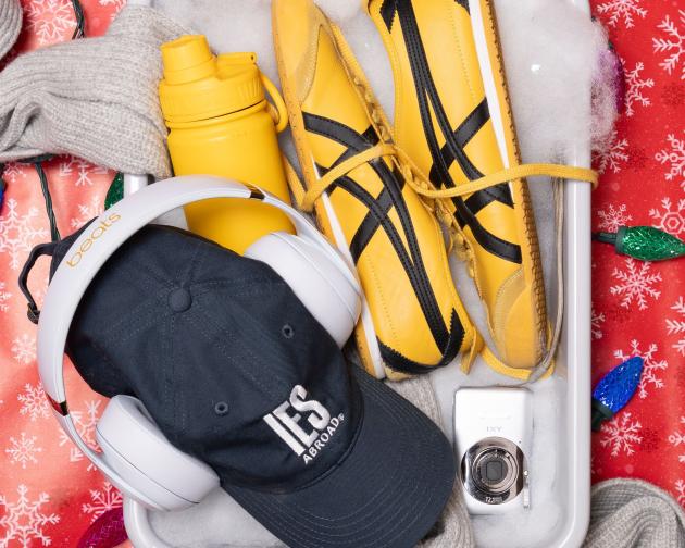 A TSA bin filled with fake snow and items for the "Jet-Setter" including an IES Abroad baseball hat, walking shoes, a water bottle, camera, headphones, and scarf