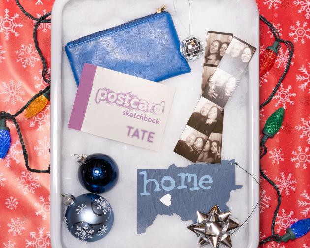 A TSA bin filled with fake snow and items for the "Wellness Enthusaist" including a cosmetic pouch, a sketchbook, an e-reader, and items from home