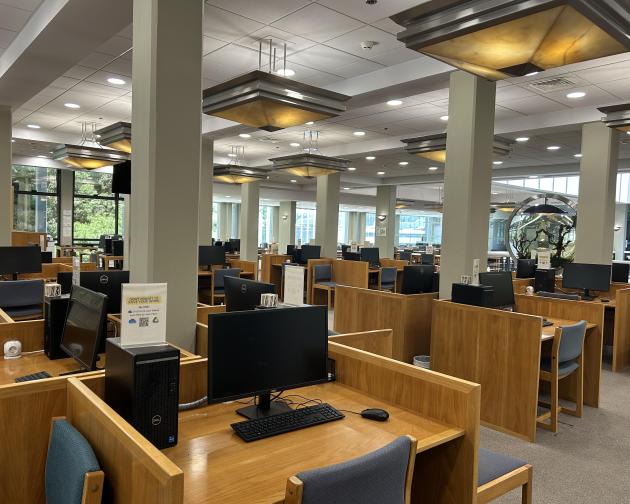 Spacious library with wooden desks and computers, large windows, and overhead lighting. This alt text was added with Al; accuracy may vary.