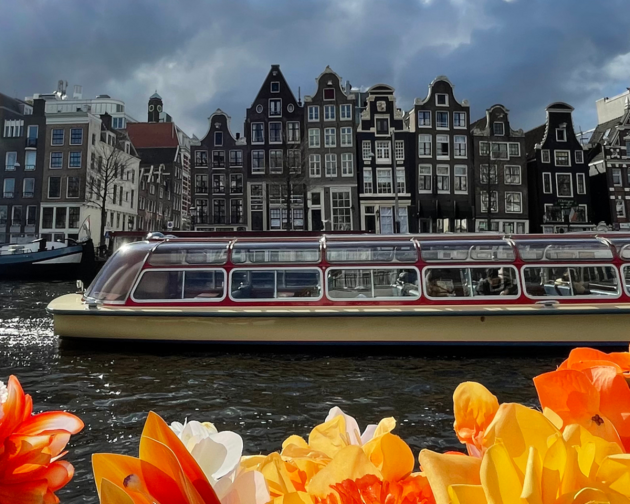 A photo of flowers in front of the water in Amsterdam, along with a boat and buildings in the background