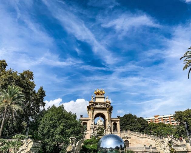 The Arc de Triomf in Barcelona, with a clear blue sky, palm trees, and a reflective sphere in front