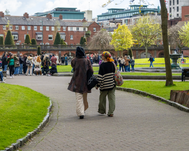 Two people walking through St. Patrick’s Park in Dublin, with a crowd gathered near a fountain and red-brick buildings in the background.
