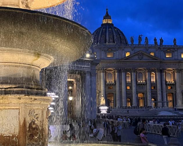 A nighttime photo of a fountain lit up in front of the Vatican.