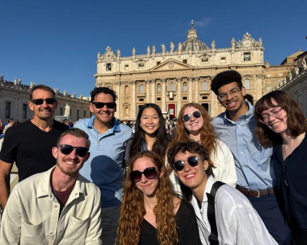 A group students wearing sunglasses and posing together on a bright, sunny day with historic architecture in the background