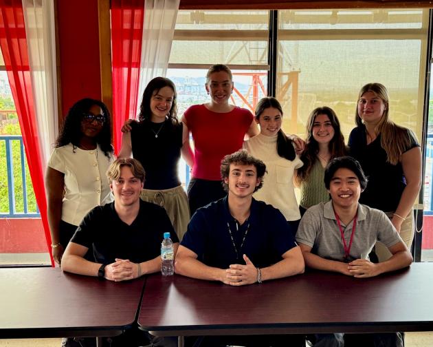A group of nine students smiling, with some sitting at a table and others standing behind them