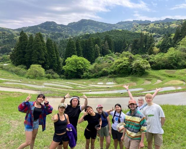 A group of diverse students smiling and posing after a hike in Japan. Green grass, trees, and mountains are seen in the distance