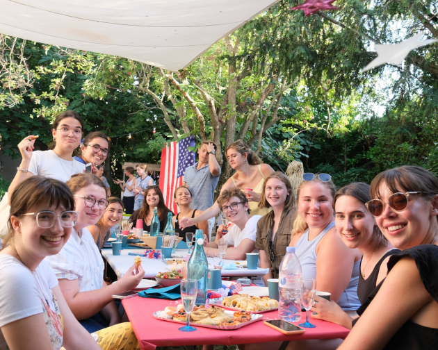 A group of students smiling around a table with an American flag hanging in the background.