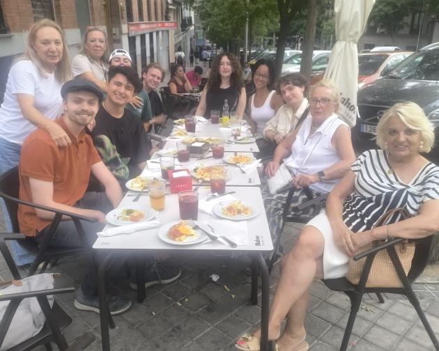 A group of eleven people sitting around a rectangular outdoor café table on a city sidewalk. They are arranged on three sides of the table, all facing toward the camera and smiling, giving a relaxed, cheerful feeling. On the left side, two older women stand just behind the seated people, leaning in to be part of the group. In front of them, a young man in an orange shirt and flat cap sits at the near left corner, turned slightly toward the center. Along the far side of the table, several younger people sit.