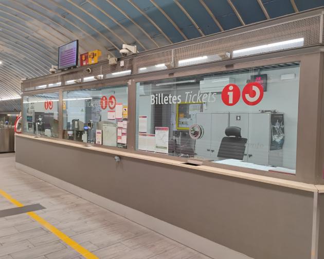 Ticket office windows in a train or metro station. A long counter runs from the foreground into the distance, with large glass windows separating the customer side from the staff area. On the glass, white text reads “Billetes Tickets” along with several large red circular symbols containing a lowercase “i” for information. Behind the glass, there are office chairs, cabinets, a computer monitor, and various posters and notices pinned up. Above the windows, electronic equipment and screens are mounted.