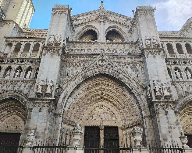 Gothic cathedral façade with a large central entrance and two side entrances, all covered in ornate stone carvings. The structure is made of pale stone and rises vertically with strong, tall pillars on the left and right sides of the main entrance. The central portal is framed by multiple pointed stone arches that recede inward, each arch ring densely filled with small sculpted figures and decorative patterns. Above the main doorway, in the tympanum area, a detailed relief scene shows many human figures.