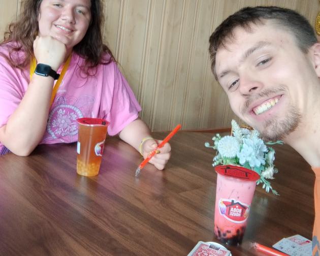 Two people sitting at a round wooden table in what looks like a cozy room with wood-paneled walls. A girl (Author’s younger sister) with light-to-medium skin and long wavy brown hair is on one side of the table, smiling toward the camera. She is wearing a pink T-shirt with a decorative design on the front, a yellow lanyard, and a dark smartwatch on her left wrist. She is resting her chin on her left hand and holding an orange spoon in her right hand over the table. In front of her is a tall orange drink cup