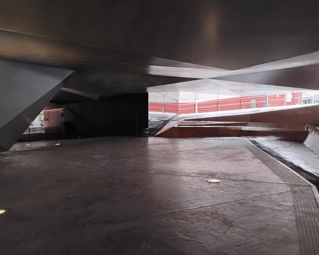 Concrete-floored underpass with angular metal ceiling panels and ramps. The image shows an interior or covered space with a smooth, slightly reflective concrete floor and several round floor lights embedded in it. The ceiling and side structures are made of large, sharp-edged metal panels forming triangular and slanted shapes, giving the space a very geometric, modern appearance. In the center-right, the floor rises in a broad ramp, with more angled surfaces leading upward toward an opening that leads outsi