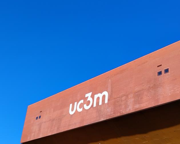 Red-brown building facade with the text “uc3m” in large white lowercase letters. The letters are centered on the wall and appear clean and modern, using a sans-serif font. The facade has a flat, rectangular shape with a slightly textured surface, as if made of brick or metal panels. Near the upper left of the wall, there are two small square windows set close together, and a similar pair of small windows appears toward the upper right. Above the building, the entire background is a clear, vivid blue sky.